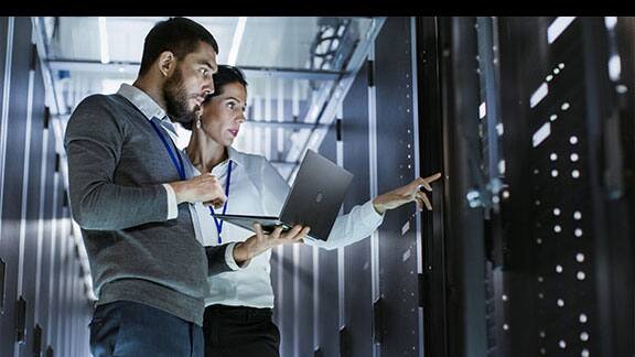 Two engineers inspecting data racks, while one holds a laptop