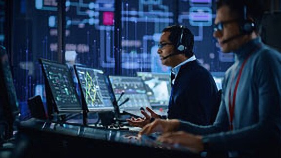 Two men wearing headsets working on computers in a control room