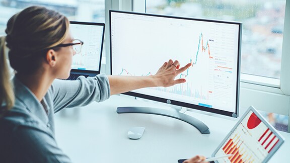 A woman pointing at a desktop monitor displaying a line graph